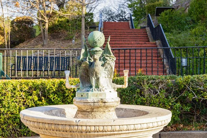Ein kunstvoller Steinbrunnen, der von Adlerskulpturen gekrönt ist, steht vor roten Treppen.
